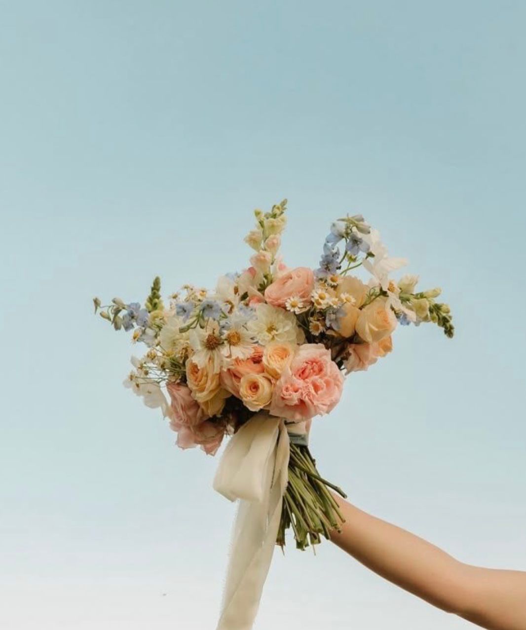A soft pastel bridal bouquet of roses, delphiniums and daisies tied with a silk ribbon, held aloft against a clear sky